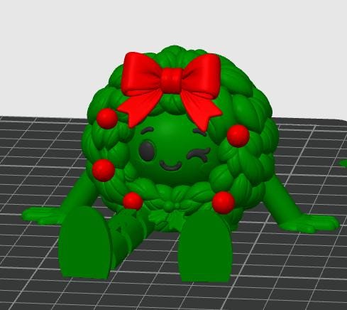May include: A green 3D-rendered character shaped like a Christmas wreath with a winking face, black eyes, and a smile. The wreath features a large red bow at the top, several small red spherical ornaments, and two green arms and legs extended outward.