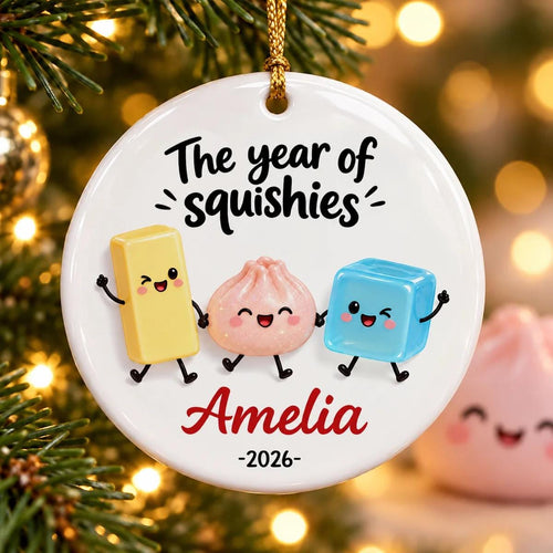 May include: A round white ceramic ornament featuring the text The year of squishies, the name Amelia, and the year 2026. The design includes three cartoon characters: a yellow rectangle, a pink dumpling, and a blue cube, all with faces.