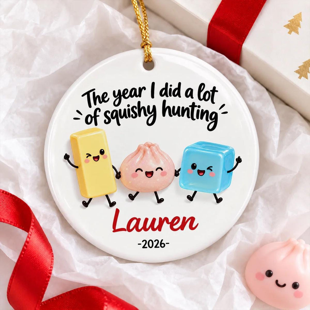 May include: A round white ceramic ornament featuring a gold hanging string. The text reads "The year I did a lot of squishy hunting" above three cartoon characters: a yellow rectangle, a pink dumpling, and a blue cube. Below are the names "Lauren" and "2026".