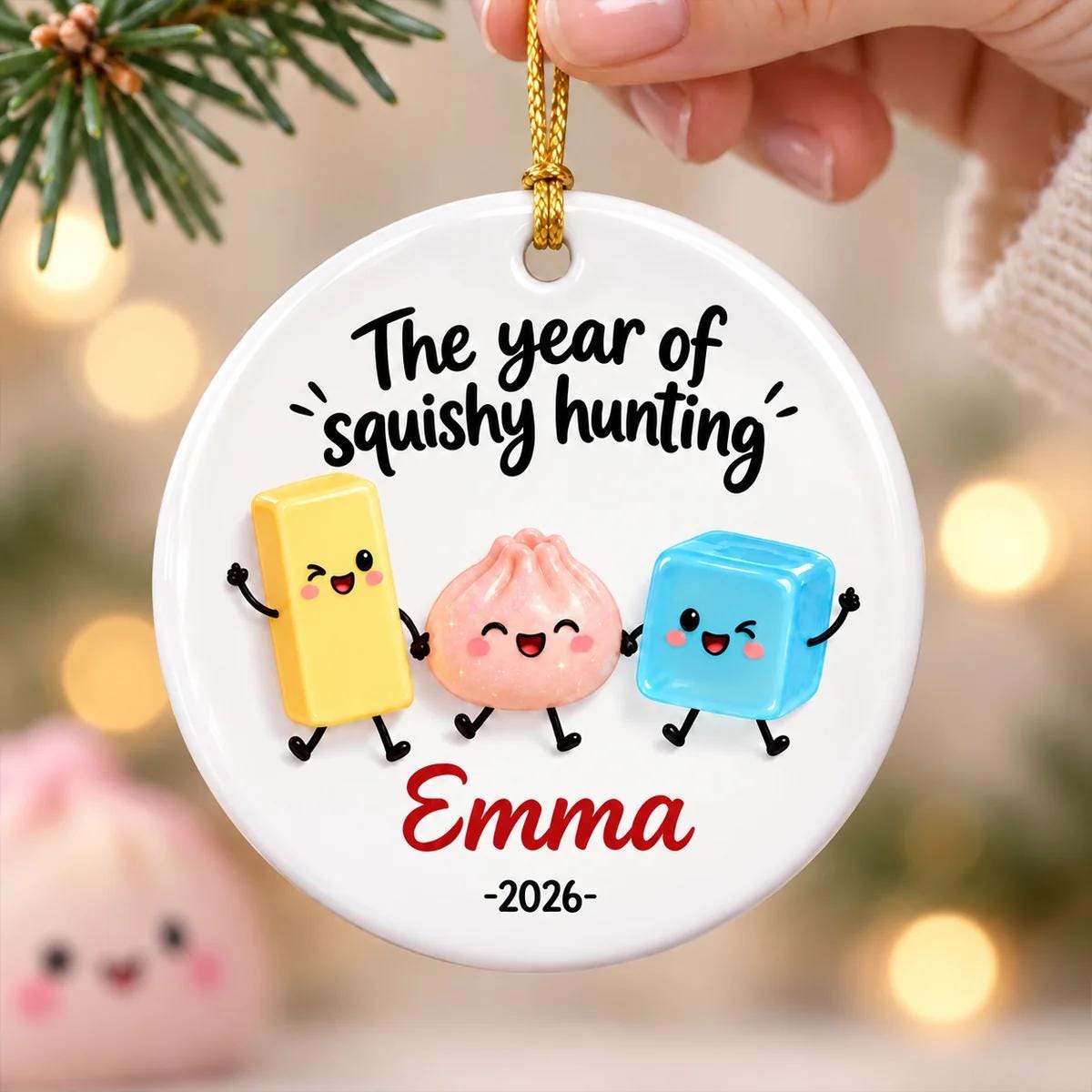 May include: A round white ceramic ornament featuring the black text The year of squishy hunting above three cartoon characters: a yellow rectangle, a pink dumpling, and a blue cube. Below them is the red name Emma and the black year 2026.
