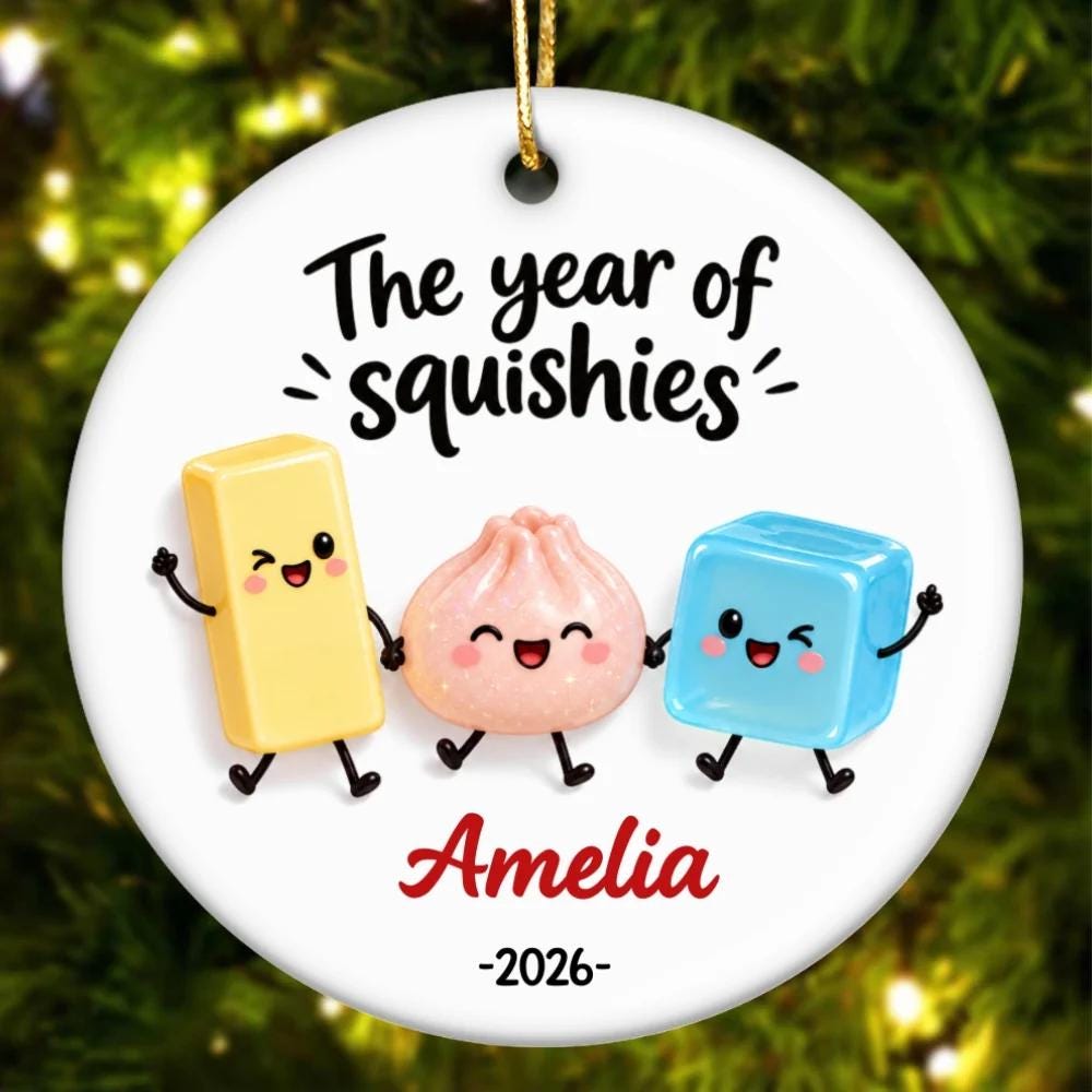 May include: A round white ceramic ornament featuring the black text The year of squishies above three cartoon characters: a yellow rectangle, a pink dumpling, and a blue cube. The name Amelia is printed in red below, with the year 2026 underneath.