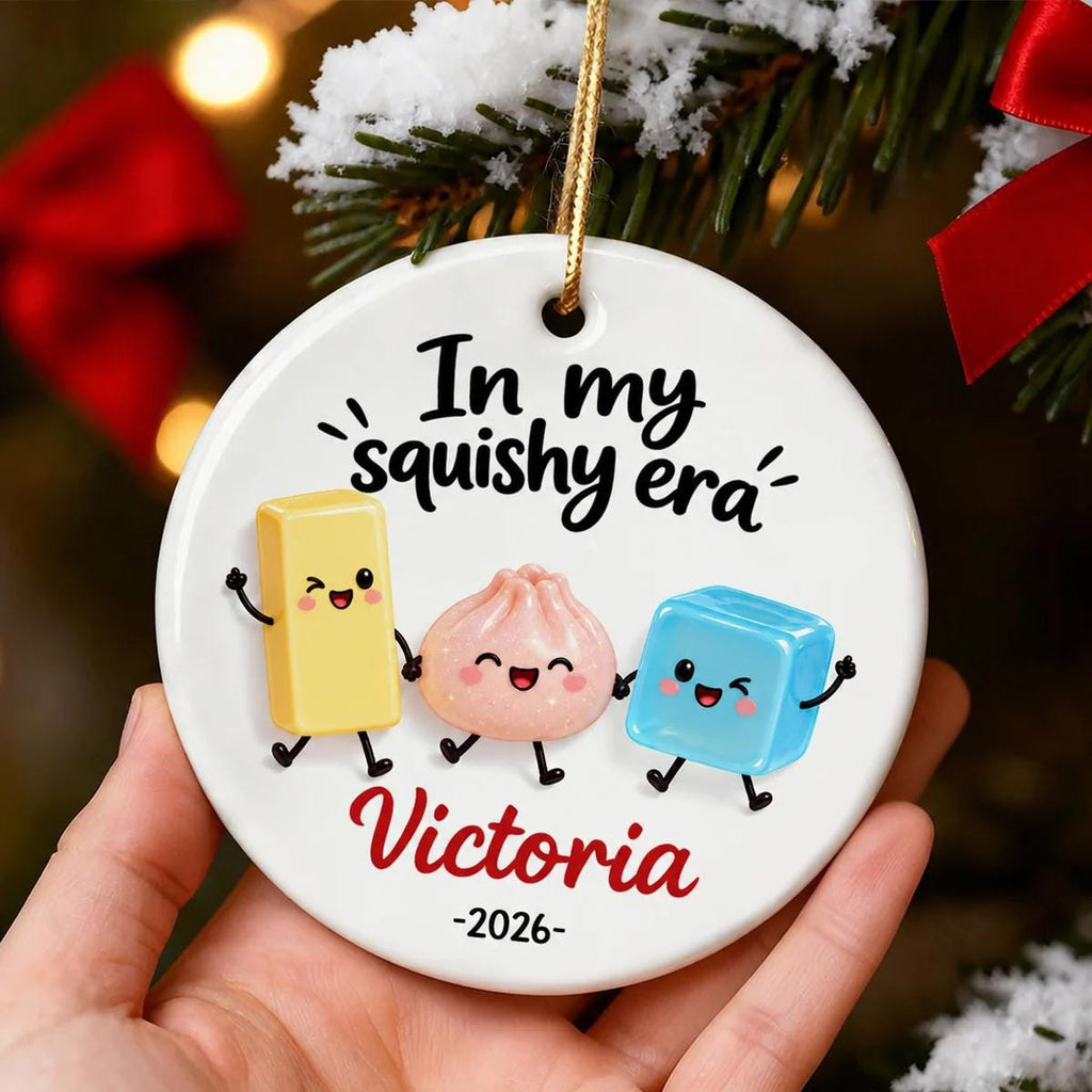 May include: A round white ceramic ornament featuring the black text In my squishy era, a yellow rectangular block, a pink dumpling, and a blue cube character with faces. The name Victoria is printed in red script below, followed by the year 2026.
