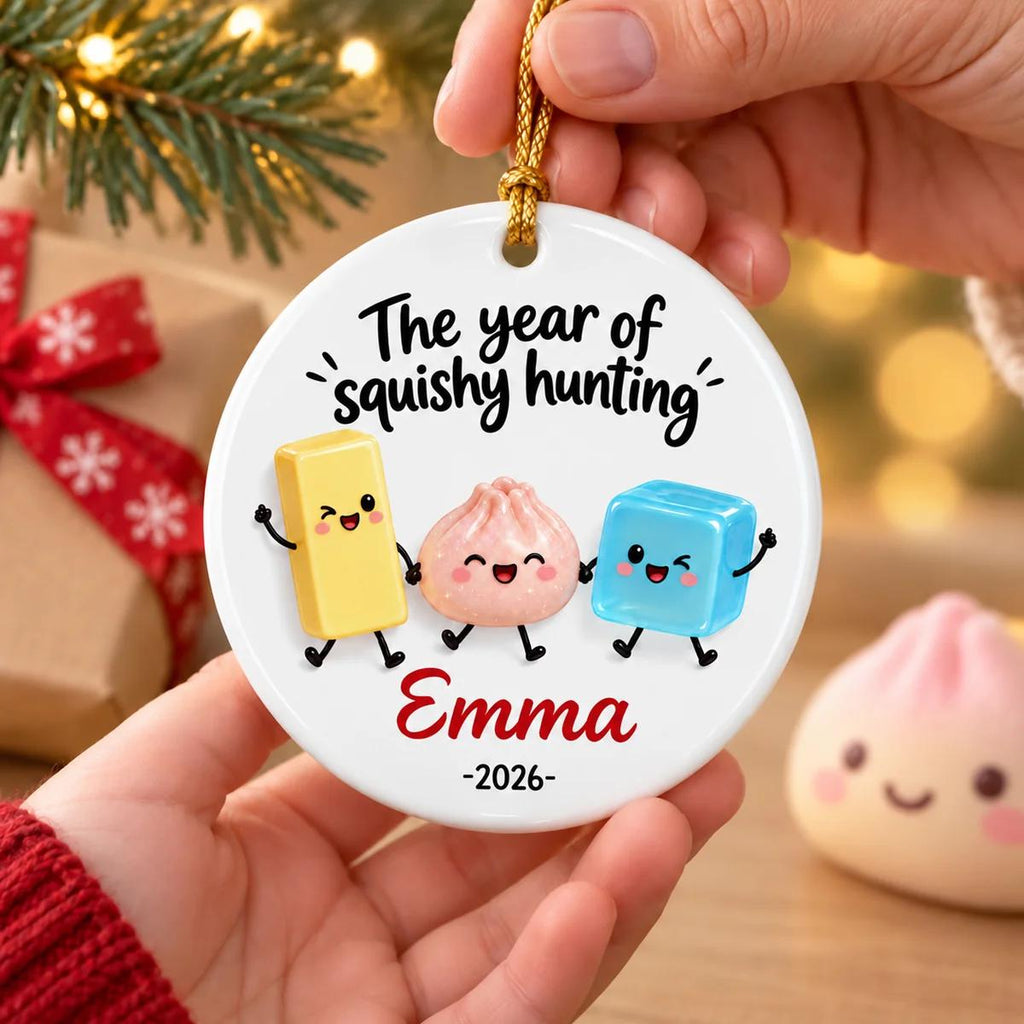 May include: A round white ceramic ornament featuring the text The year of squishy hunting, the name Emma, and the year 2026. The design includes three cartoon characters: a yellow rectangle, a pink dumpling, and a blue cube, all with faces.