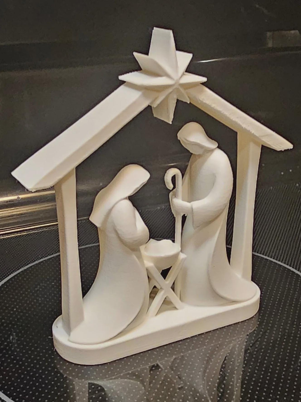 May include: A white 3D printed nativity scene figurine featuring a stylized stable structure with a star at the peak. The scene depicts two figures standing on either side of a small manger, with one figure holding a staff, all mounted on a base.
