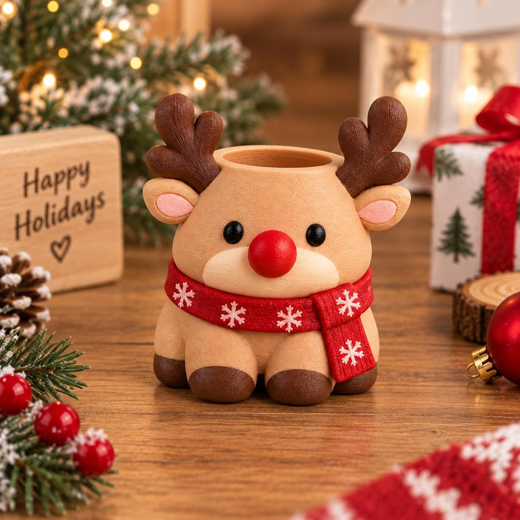May include: A tan reindeer-shaped planter or desk organizer featuring dark brown antlers, black eyes, a bright red nose, and a red scarf with white snowflake patterns wrapped around its neck. The figure has a hollow opening at the top.