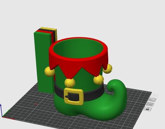 May include: A green boot-shaped container featuring a red scalloped cuff with yellow jingle bells, a black belt with a yellow buckle, and a tall green rectangular attachment with stacked red, yellow, and black layers at the top edge.