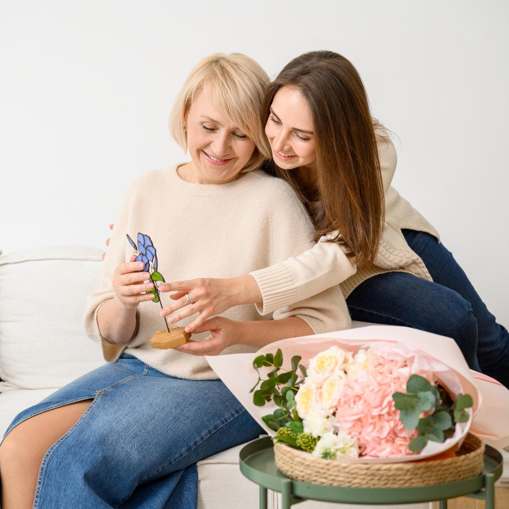 May include: A stained glass flower ornament featuring blue petals and green leaves, mounted on a small wooden base. The flower is held by two individuals, one wearing a beige sweater and blue denim skirt, the other in a beige sweater and blue jeans.