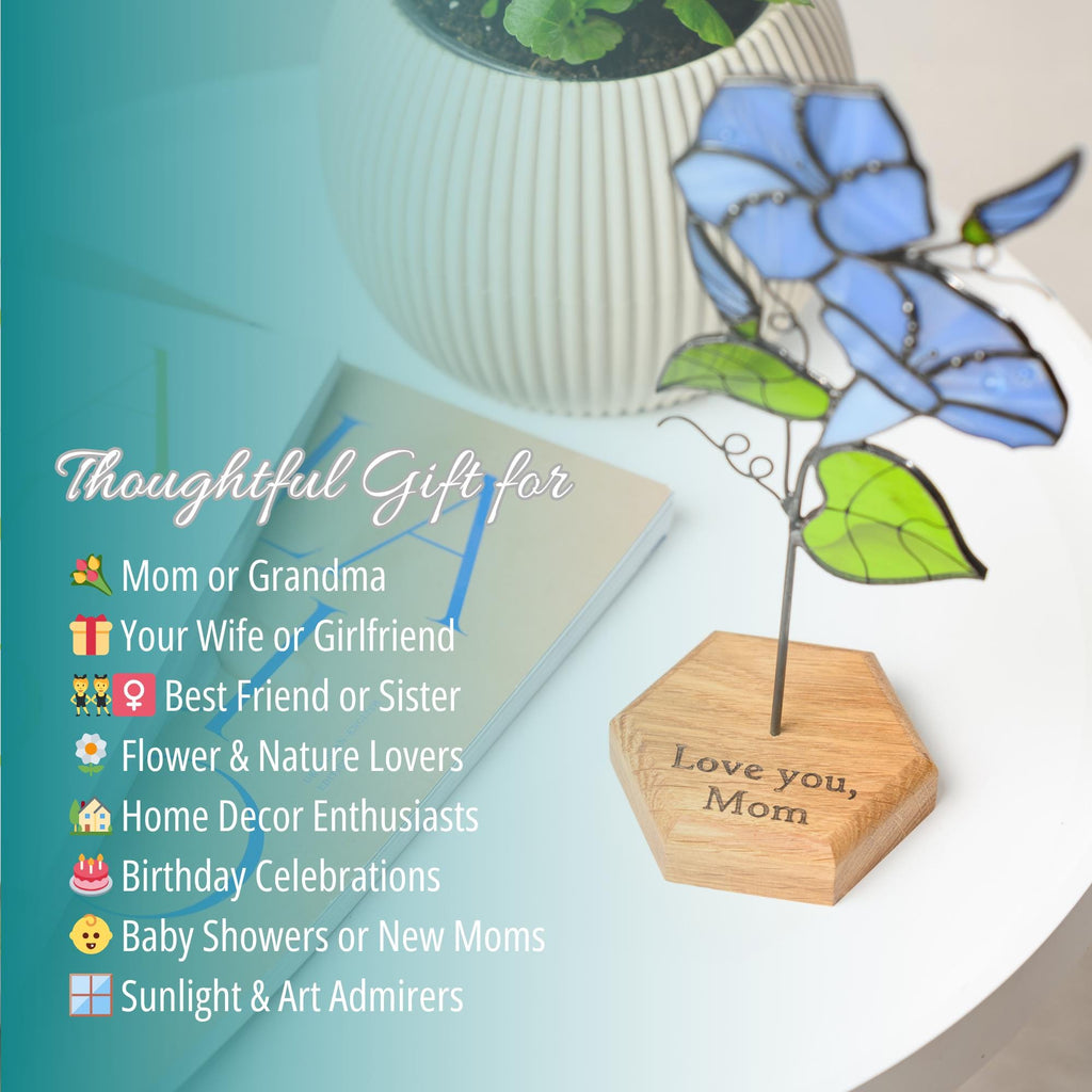 May include: A stained glass flower sculpture with light blue petals and green leaves on a thin metal stem. The sculpture is mounted on a hexagonal wooden base engraved with the text "Love you, Mom". The base sits on a white surface.