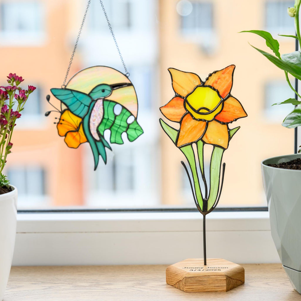 May include: A stained glass hummingbird suncatcher with a circular background and a stained glass daffodil flower on a wooden base. The wooden base features the engraved text "Jimmy Janson 3/4/2025" and the pieces are displayed near a window.