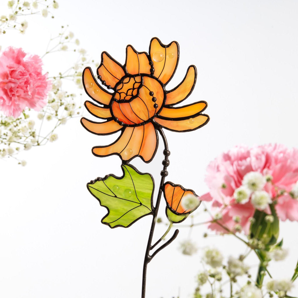May include: A stained glass flower ornament featuring a large orange bloom with dark solder lines, a single light green leaf, and a small orange bud on a thin metal stem. The glass pieces have a smooth, translucent finish with visible texture.