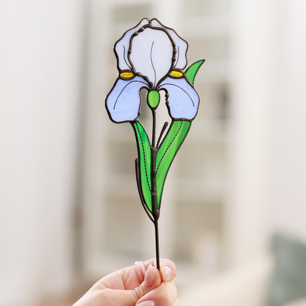 May include: A stained glass iris flower ornament featuring white and light blue petals with yellow accents. The flower has a green stem and leaves, all outlined in dark metal solder. The piece is crafted with individual glass segments.