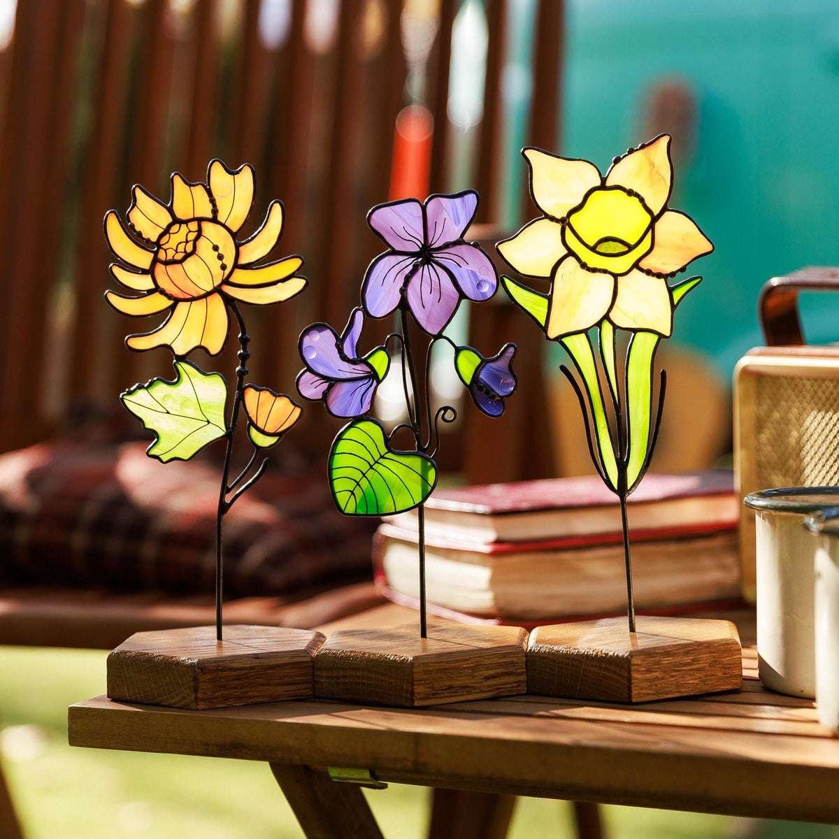 May include: Three stained glass flower sculptures on hexagonal wooden bases. The left features a yellow chrysanthemum, the center a purple violet, and the right a yellow daffodil. Each flower has green leaves and thin metal stems.