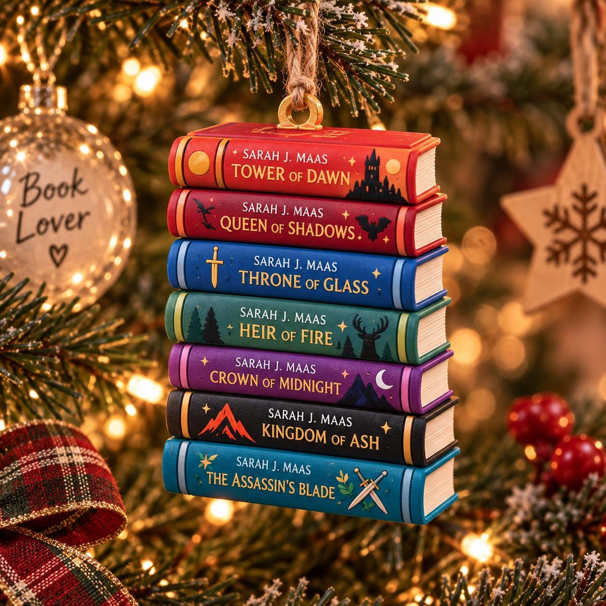 May include: A hanging Christmas tree ornament featuring a stack of seven miniature books by Sarah J. Maas. The spines are colored red, dark red, blue, green, purple, black, and teal, each displaying its title, author name, and unique graphic icons.