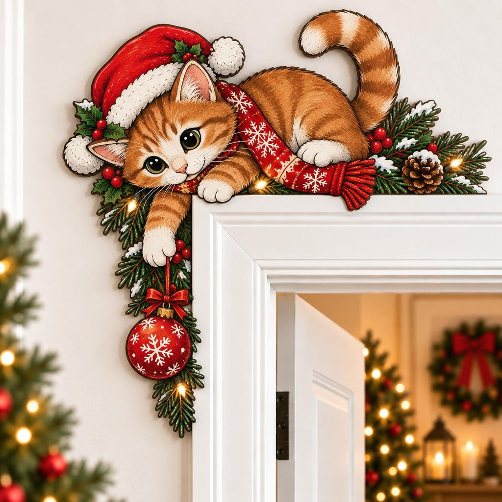 May include: A decorative wooden door corner topper featuring an orange tabby kitten wearing a red Santa hat and a red scarf with white snowflake patterns. The kitten is nestled among green pine branches, red berries, and a hanging red ornament.