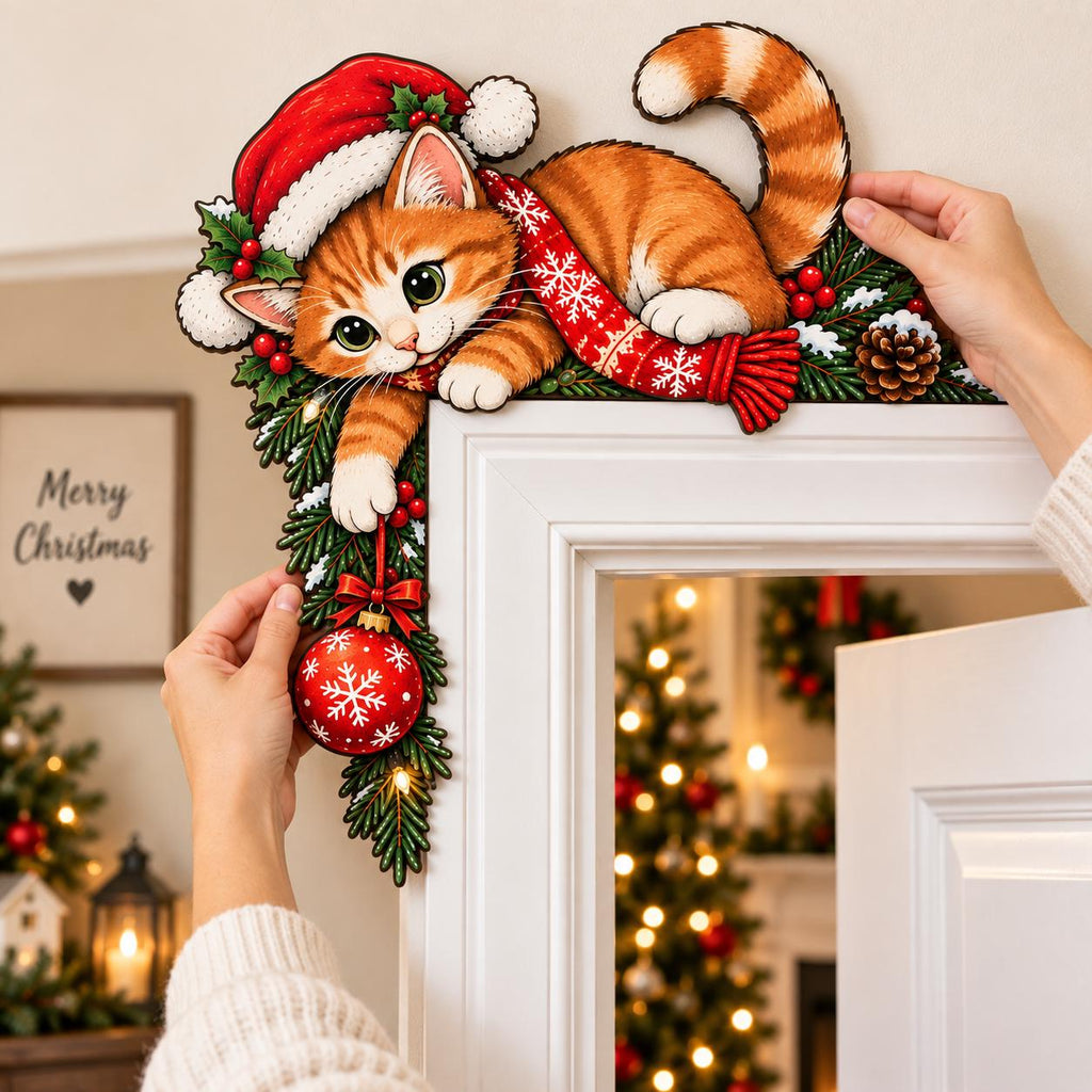 May include: A decorative wooden door corner topper featuring an orange tabby kitten wearing a red Santa hat and a red scarf with white snowflake patterns. The kitten is nestled among green pine branches, red berries, and a pinecone, holding a red ornament.