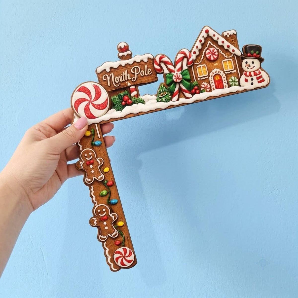 May include: An L-shaped wooden door corner decoration featuring a gingerbread house, a snowman, candy canes, and peppermint candies. The sign reads North Pole in white script. Two gingerbread cookies are attached to the vertical side.