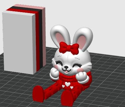 May include: A white rabbit figurine with long ears, pink inner ears, and a red bow on its head. The rabbit has a smiling expression, pink cheeks, and is wearing a red outfit with white heart accents. Beside it stands a rectangular block with layers.