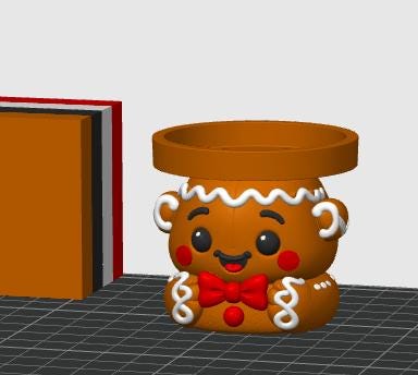May include: A brown gingerbread person figurine with a circular top opening, featuring white icing details, black eyes, red cheeks, and a red bow tie. The figure has a smiling expression and is positioned next to a stack of colored rectangular blocks.