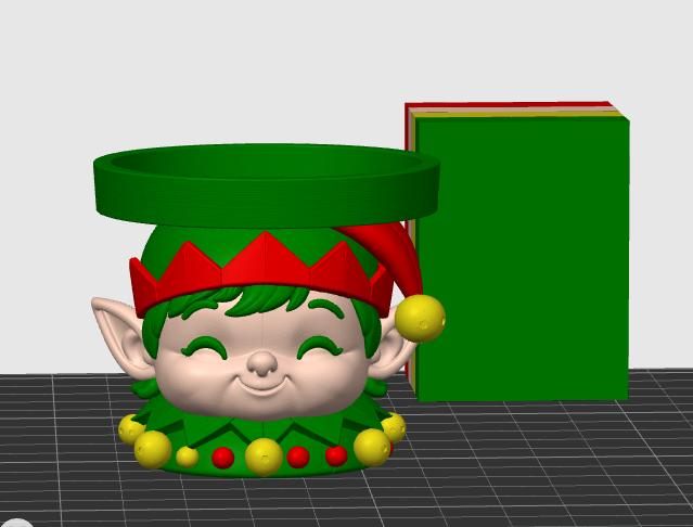 May include: A 3D rendered elf-themed bowl featuring a smiling face with pointed ears, a red hat with a yellow pom-pom, and a green collar adorned with yellow and red spheres. The bowl has a wide green rim and sits next to a green rectangular block.