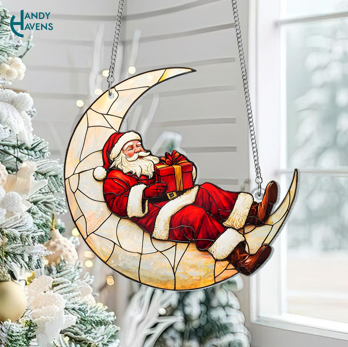 May include: A stained-glass style Christmas decoration featuring Santa Claus reclining on a crescent moon. Santa wears a red suit with white trim and holds a wrapped gift. The moon is a light beige color with a stained-glass effect, suspended by silver chains.