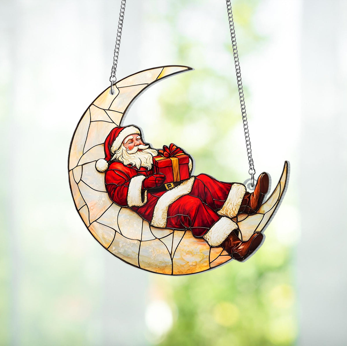 May include: A stained glass Christmas decoration featuring Santa Claus reclining on a crescent moon. Santa is dressed in a red suit with white trim and holding a wrapped gift. The moon is cream-colored with a geometric pattern, suspended by silver chains.