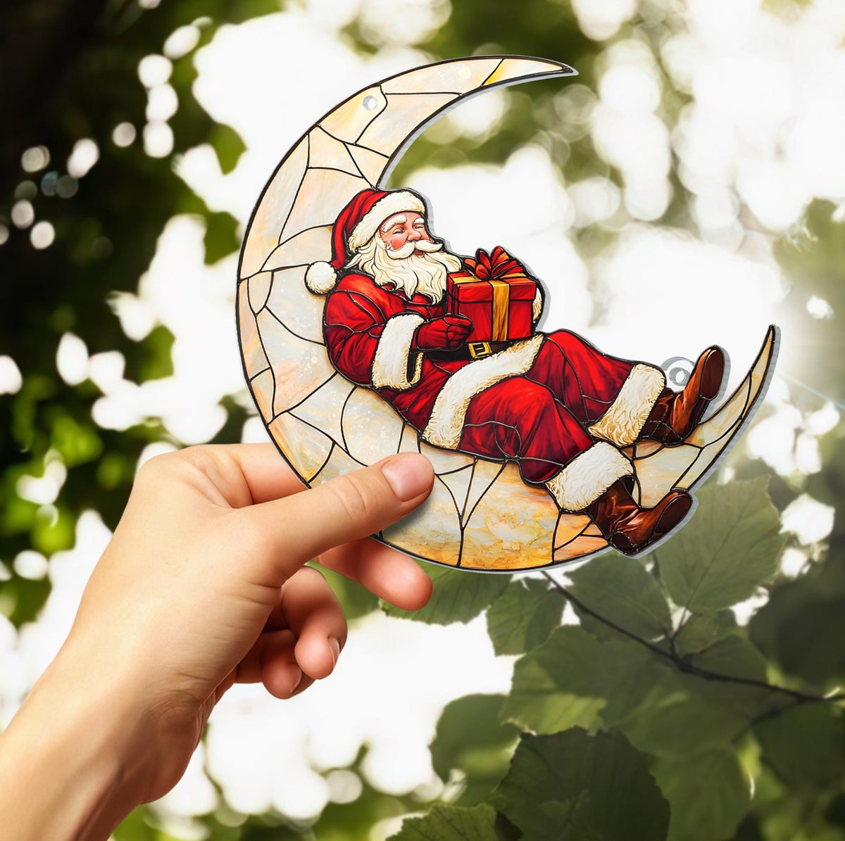 May include: A stained glass Christmas ornament featuring Santa Claus reclining on a crescent moon. Santa is dressed in a red suit with white trim, holding a wrapped gift. The moon is a light beige color with a stained glass effect.