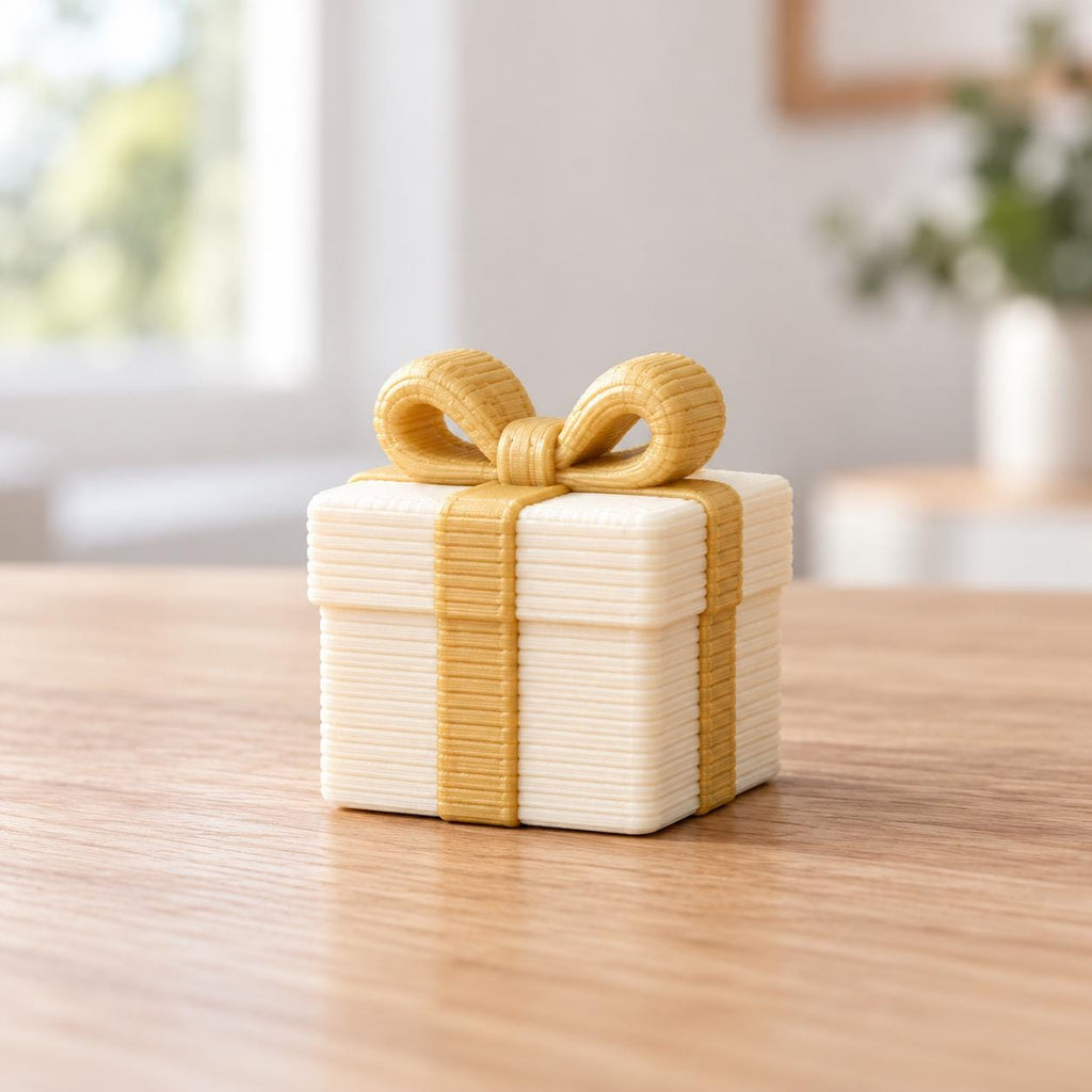 May include: A small, square, white gift box figurine featuring a textured, layered surface. The box is wrapped with a gold-colored ribbon that extends vertically around the sides and forms a prominent, detailed bow on the top center.