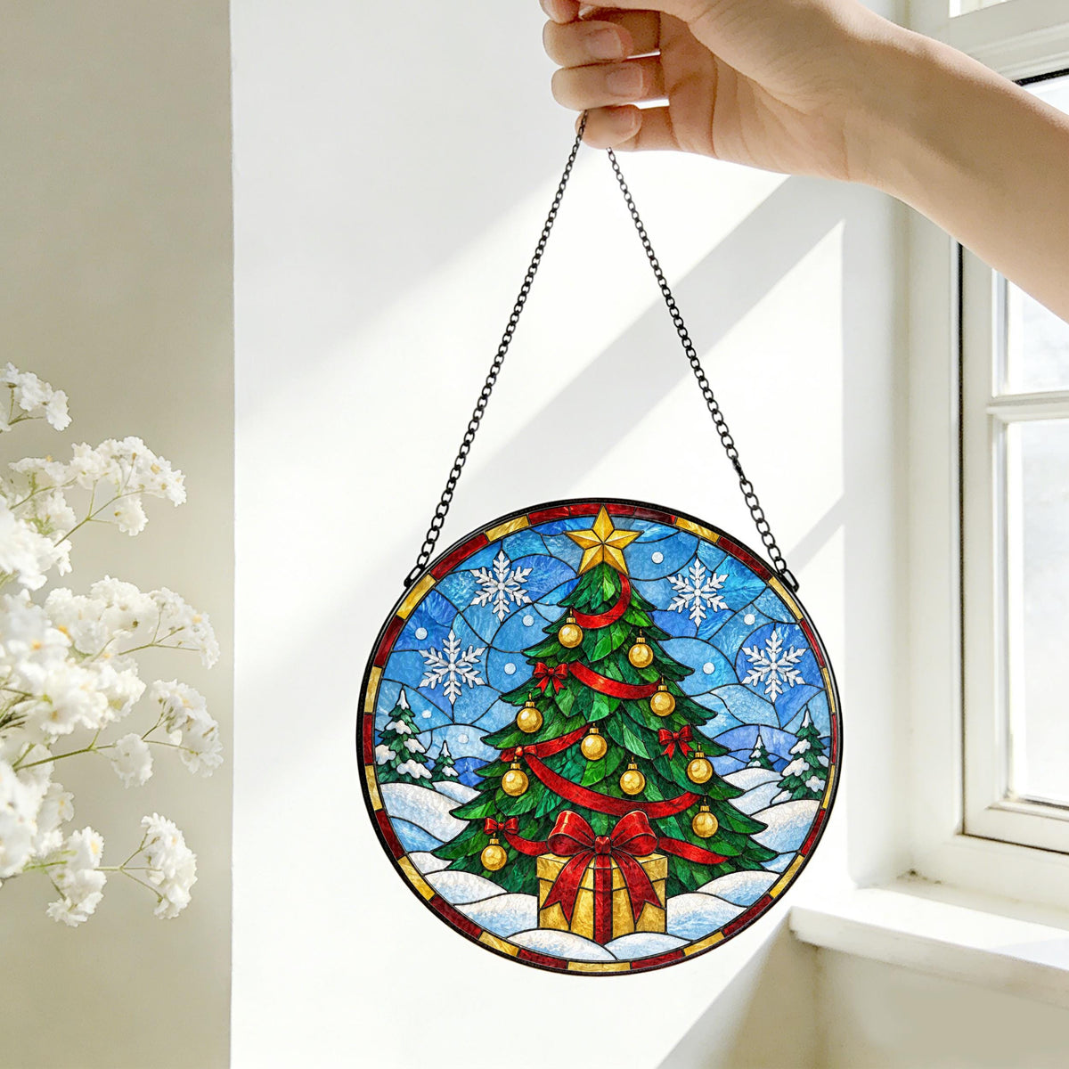 May include: A circular stained glass suncatcher featuring a green Christmas tree decorated with gold ornaments, a red ribbon, and a yellow star topper, set against a blue snowy background with white snowflakes and a gift box at the base.
