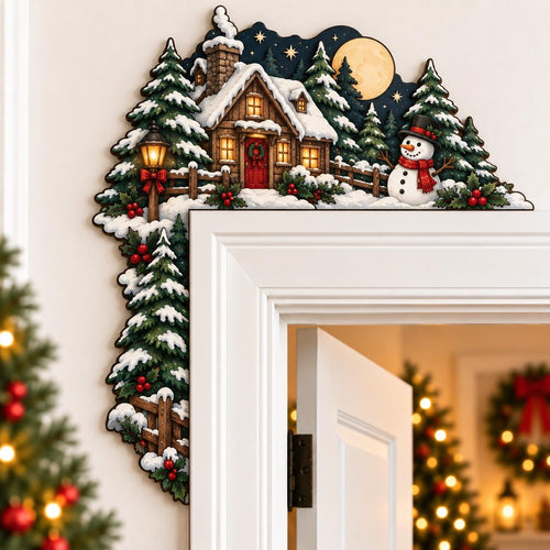 May include: A decorative wooden door corner topper featuring a winter scene with a snow-covered cabin, a snowman wearing a black top hat and red scarf, evergreen trees, a glowing street lamp, and a full moon against a dark blue night sky.