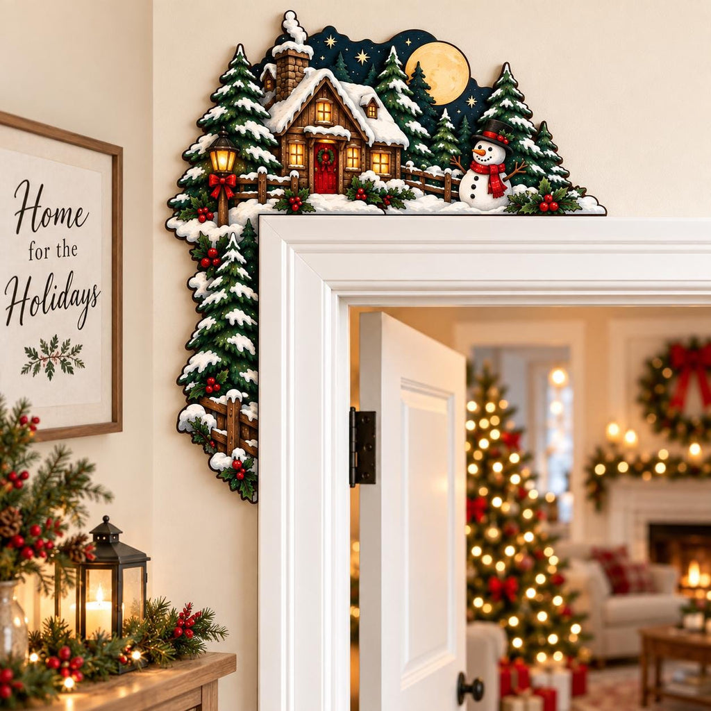 May include: A decorative wooden door corner topper featuring a winter scene with a snow-covered cabin, evergreen trees, a snowman, and a full moon against a dark blue night sky. The design includes a lit lantern and festive holly accents.