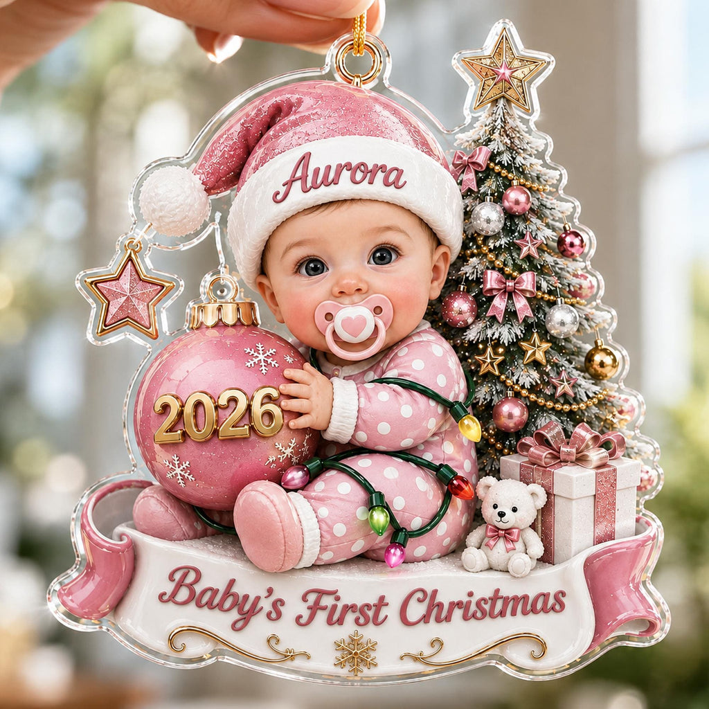 May include: A clear acrylic Christmas ornament featuring a baby in pink polka dot pajamas and a matching hat with the name Aurora. The baby holds a pink ornament labeled 2026. Beside them is a decorated Christmas tree, a teddy bear, and a gift box.