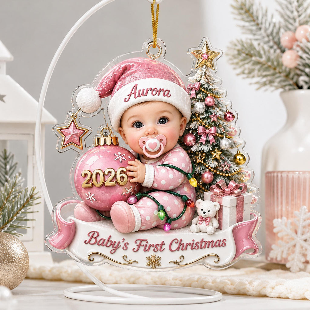 May include: A decorative acrylic Christmas ornament featuring a baby wearing a pink hat and polka dot pajamas, holding a pink ornament labeled 2026. The design includes a Christmas tree, a teddy bear, a gift box, and a banner reading Baby's First Christmas.