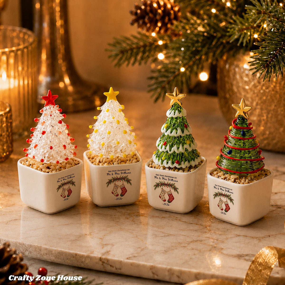 May include: Four miniature Christmas tree ornaments in square white ceramic pots. Each tree is topped with a gold or red star and sits on a bed of small pebbles. The pots feature a printed illustration of two stockings and the text Our First Christmas.
