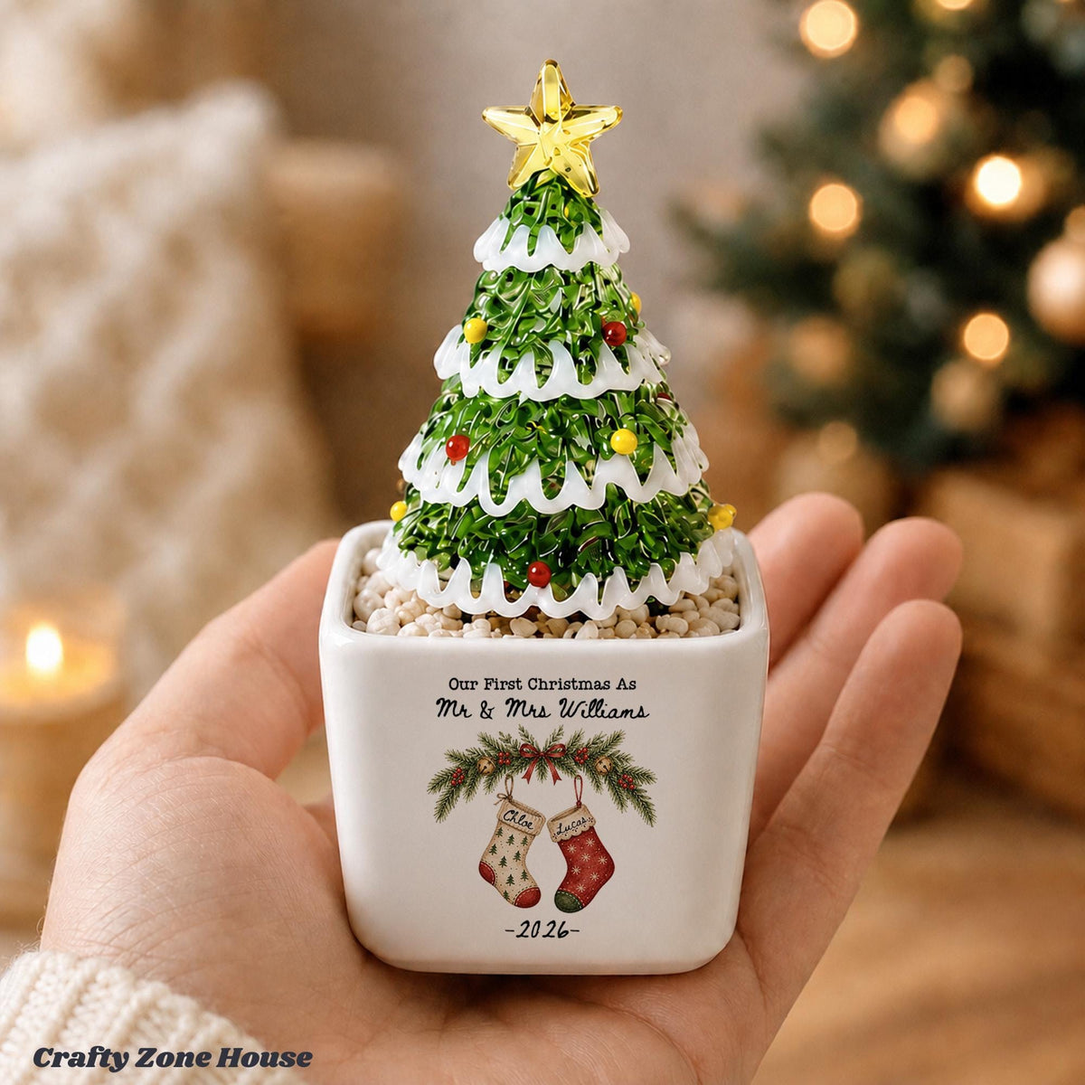 May include: A small decorative Christmas tree ornament in a white square ceramic planter. The tree is green with white snow-like trim, colorful ornaments, and a yellow star topper. The planter features the text Our First Christmas As Mr & Mrs Williams, a graphic of two stockings, and the year 2026.