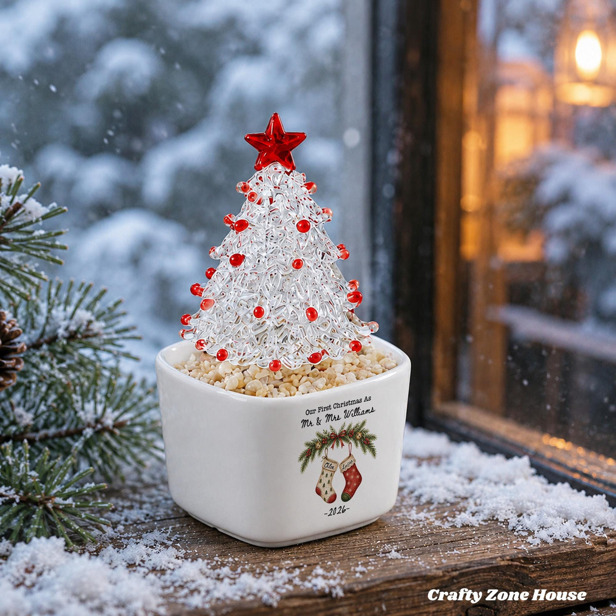 May include: A clear glass Christmas tree ornament with red glass beads and a red star topper, set in a white square ceramic planter filled with small stones. The planter features the text Our First Christmas As Mr & Mrs Williams 2026.
