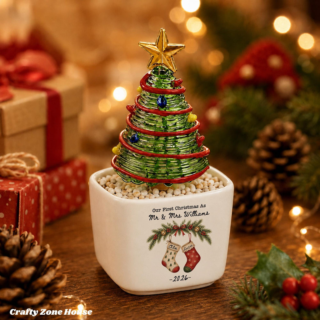 May include: A decorative tabletop Christmas tree made of green wire wrapped with red ribbon and small colored beads, topped with a gold star. It sits in a white square ceramic planter featuring the text Our First Christmas As Mr & Mrs Williams, a festive illustration of two stockings, and the year 2026.