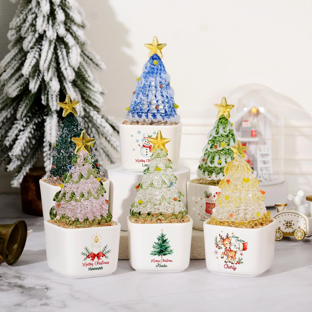 May include: A collection of six small, decorative Christmas tree ornaments in white ceramic pots. Each tree is made of textured, translucent material in various colors, topped with a gold star. The pots feature festive graphics and names.