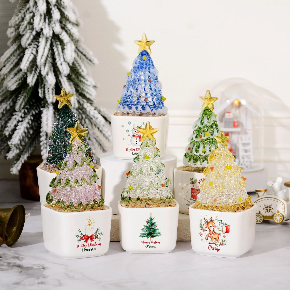 May include: A collection of six small, decorative Christmas tree ornaments in white ceramic pots. Each tree is made of textured, translucent material in various colors, topped with a gold star. The pots feature festive graphics and names.