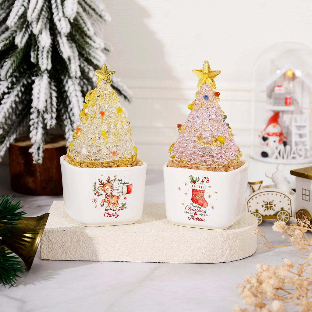 May include: Two decorative tabletop Christmas trees with textured, translucent yellow and pink glass foliage topped with gold stars. Each tree sits in a white square ceramic planter featuring festive illustrations and the names Cherly and Marcia.