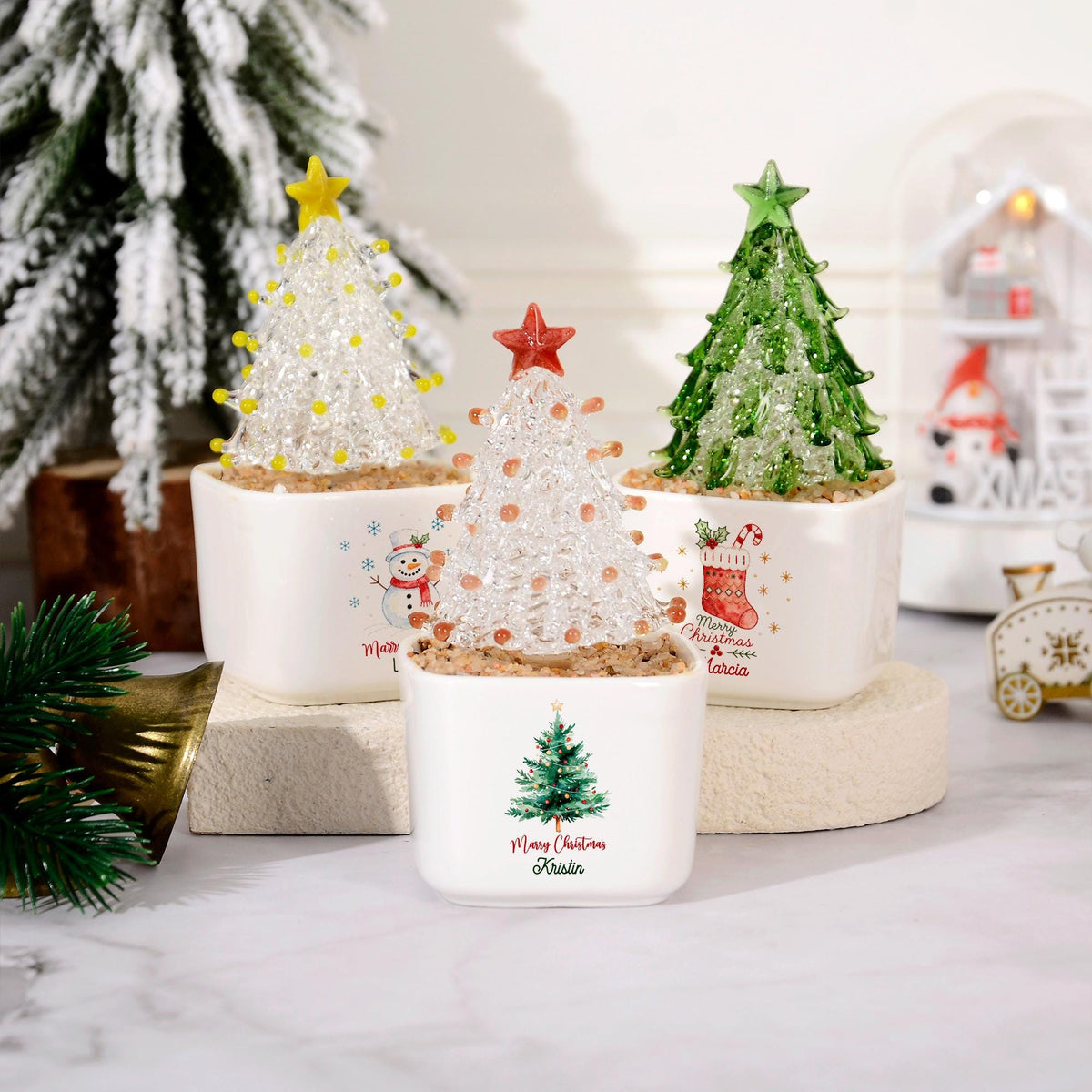 May include: Three small glass Christmas tree ornaments in white square ceramic pots. The trees are clear, green, and white with colored stars and beads. The pots feature printed holiday graphics of a snowman, a stocking, and a tree with names.