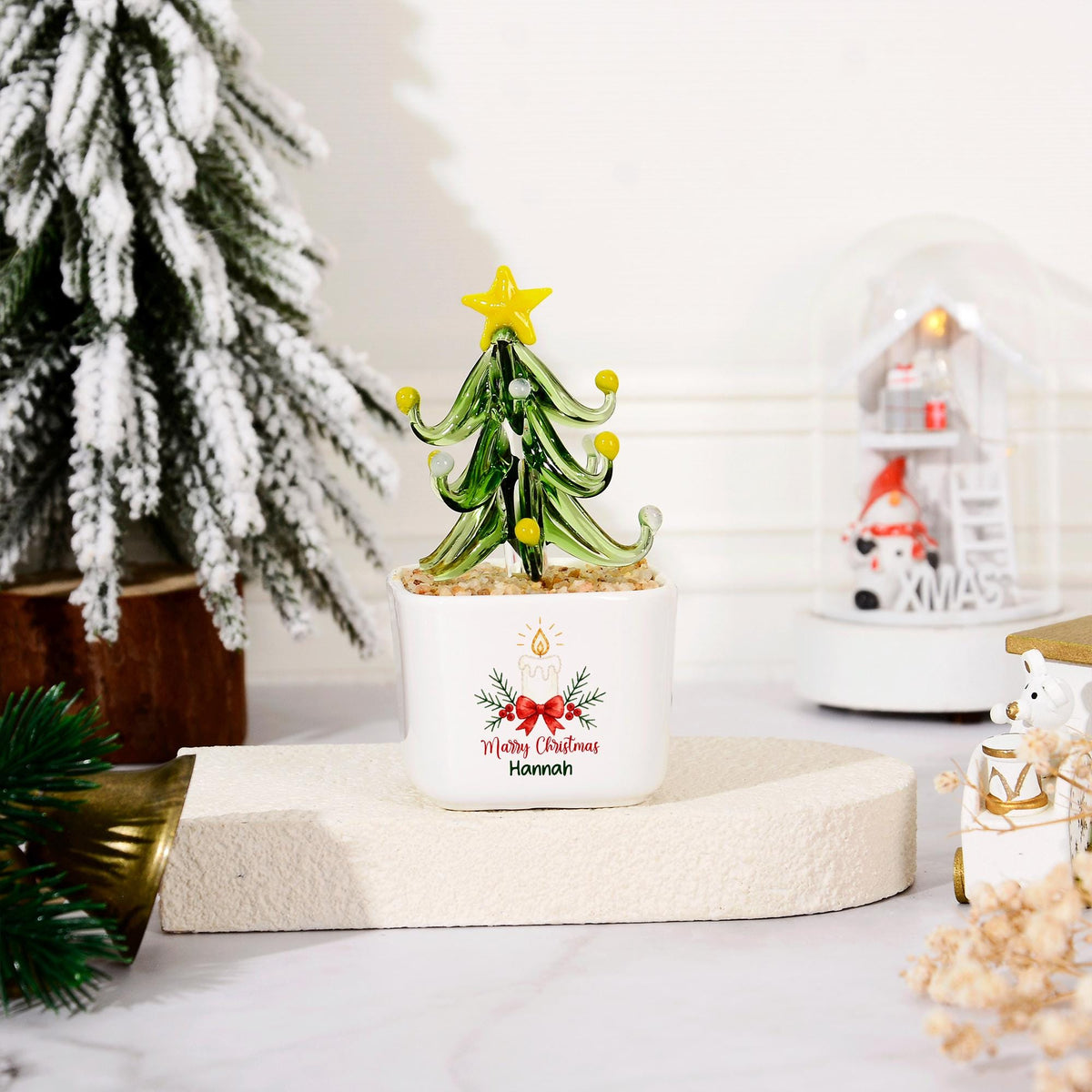 May include: A glass Christmas tree ornament with green branches, yellow accents, and a yellow star topper, set in a white square planter. The planter features a printed graphic of a candle, red bow, and holly with the text Merry Christmas Hannah.