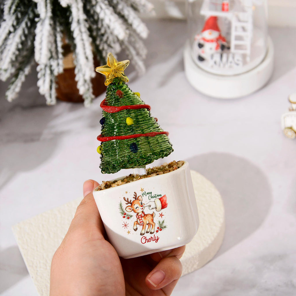 May include: A small white ceramic planter containing a green glass Christmas tree ornament with a yellow star topper, red garland, and colorful baubles. The planter features a printed graphic of a reindeer, a hand, and the text Merry Christmas Cherly.