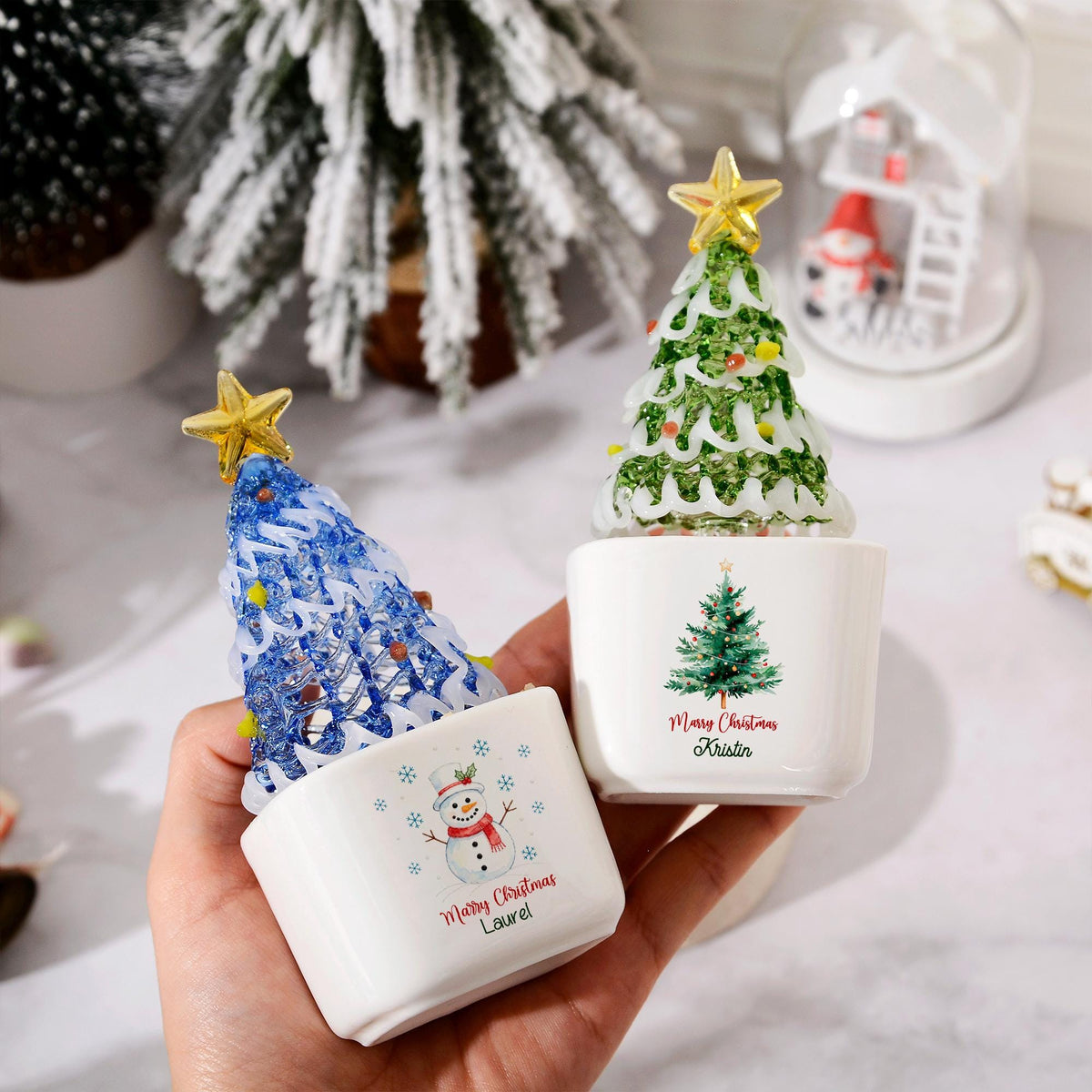 May include: Two white ceramic square planters featuring glass Christmas tree ornaments with gold stars on top. One planter displays a snowman graphic with the text Merry Christmas Laurel, while the other shows a pine tree with Merry Christmas Kristin.