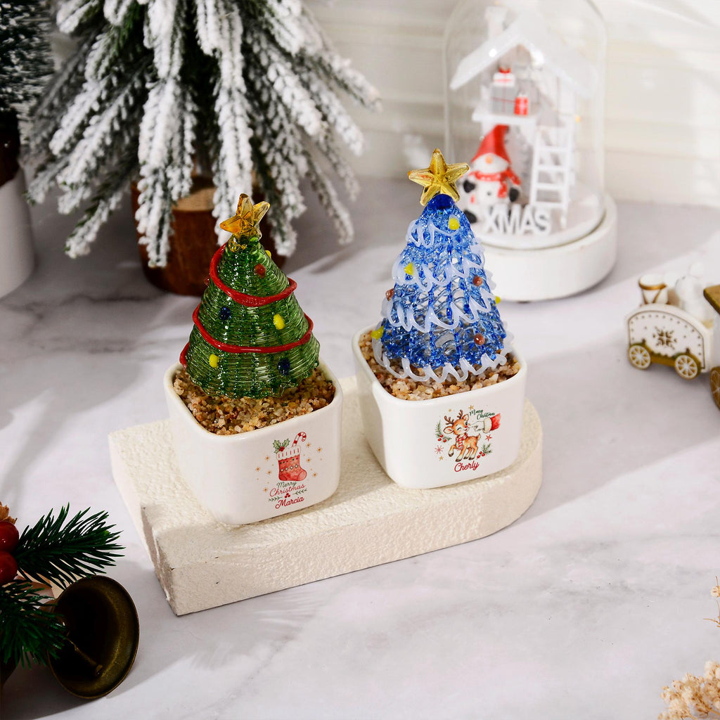 May include: Two miniature Christmas tree ornaments in square white ceramic pots. One tree is green with red garland and yellow dots, the other is blue with white swirls and blue dots. Both feature a gold star topper and a festive graphic print.