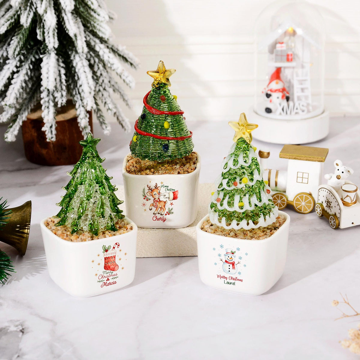 May include: Three miniature glass Christmas trees in square white ceramic pots. Each tree is topped with a gold star and sits on a bed of small pebbles. The pots feature festive illustrations of a reindeer, a stocking, and a snowman with names.