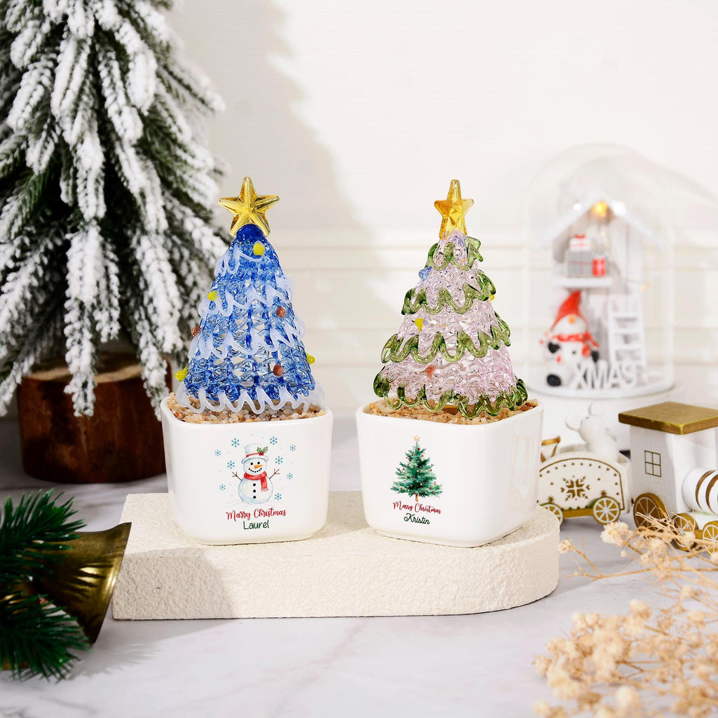 May include: Two small decorative Christmas trees in white square ceramic pots. One tree is blue with a gold star, the other is light pink with green accents and a gold star. The pots feature printed snowmen, trees, and the text Merry Christmas.