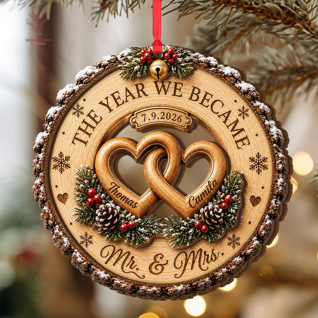 May include: A round wooden Christmas ornament with bark edges and snow accents. It features two interlocking hearts engraved with the names Thomas and Camila, the text THE YEAR WE BECAME, 7.9.2026, and Mr. & Mrs. It is decorated with pine needles, pinecones, red berries, and a gold bell.