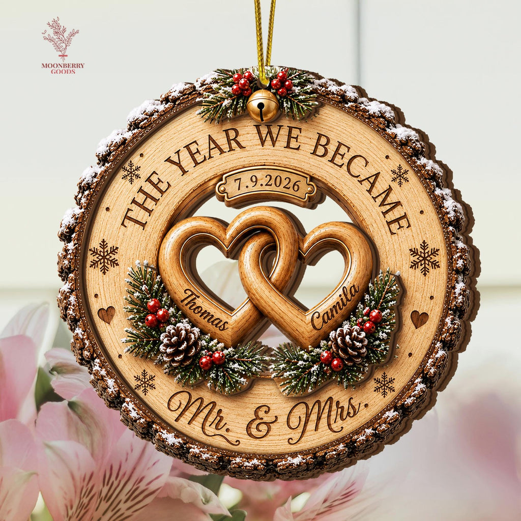 May include: A round wooden ornament with a bark-textured edge, featuring two interlocking hearts engraved with the names Thomas and Camila. The text reads THE YEAR WE BECAME, 7.9.2026, and Mr. & Mrs., accented with pine branches, red berries, and snowflakes.