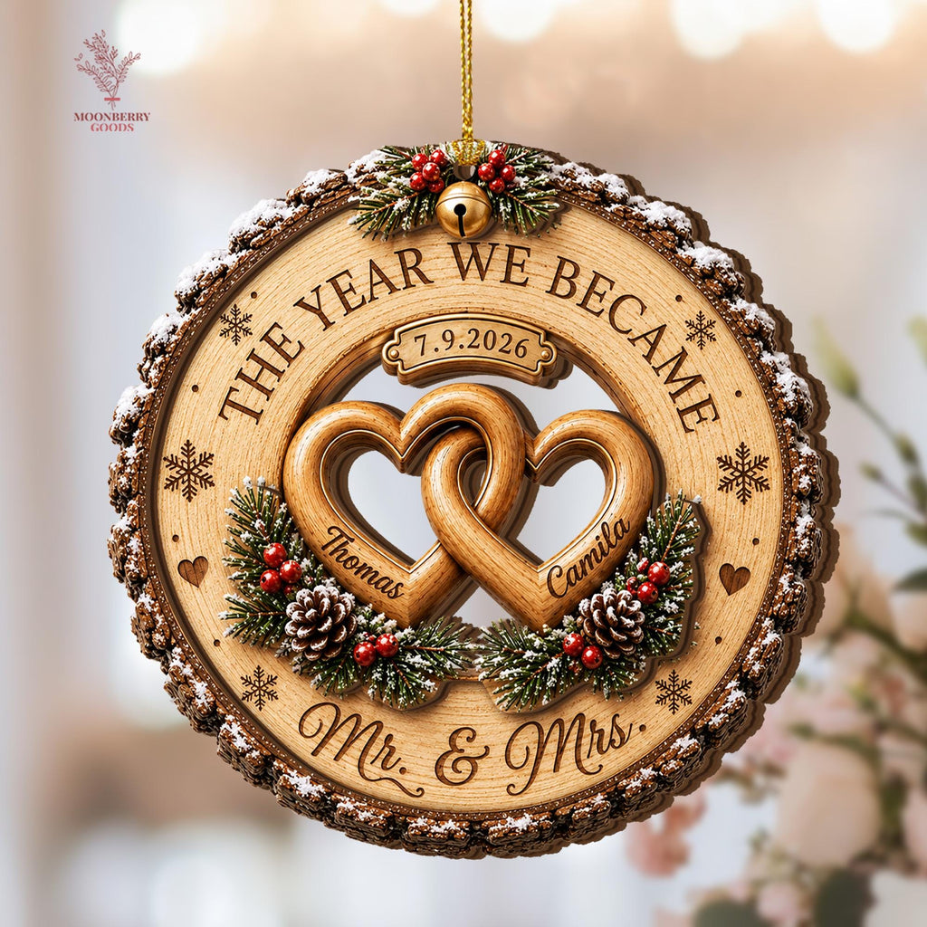 May include: A circular wooden ornament with bark edges, featuring two interlocking hearts engraved with the names Thomas and Camila. The text reads THE YEAR WE BECAME, 7.9.2026, and Mr. & Mrs. It is decorated with pine branches, red berries, and a gold bell.