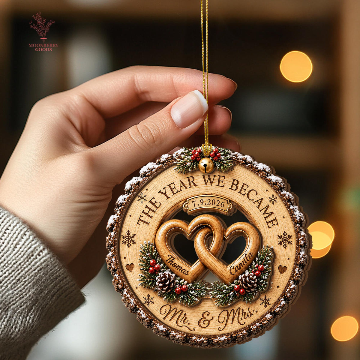 May include: A round wooden Christmas ornament featuring two interlocking heart cutouts, engraved with the names Thomas and Camila, the date 7.9.2026, and the text THE YEAR WE BECAME MR. & MRS. It is decorated with pine branches, red berries, and a gold bell.